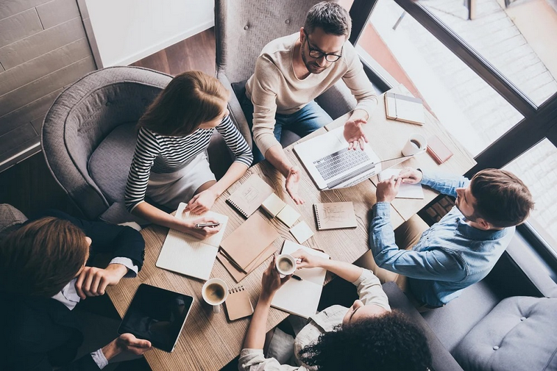 A group of four diverse professionals collaboratively working with laptops and documents around a small table in a cozy office setting.