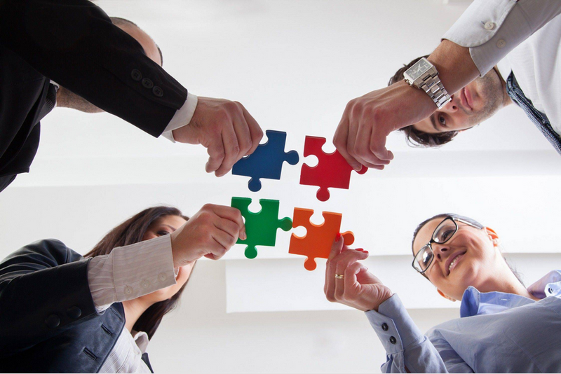 Four people collaboratively connecting puzzle pieces from below, symbolizing teamwork and problem-solving.