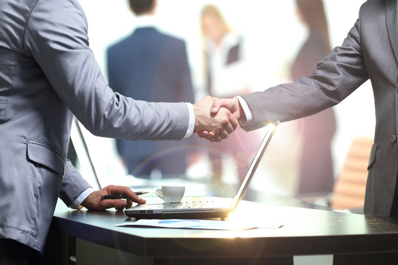 Two businessmen shaking hands over a table with a laptop in a professional office setting.